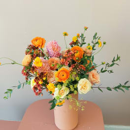 Mixed bouquet of orange, pink, and cream flowers in a blush vase
