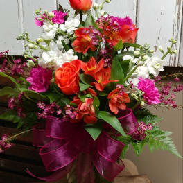 Mixed bouquet of red, orange, white, and pink flowers in a glass vase with a magenta ribbon