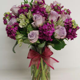 Round arrangement of lavender roses and bright purple flowers in a clear vase with a pink bow