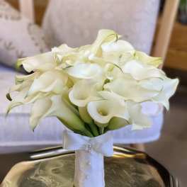 Hand-tied bouquet of white calla lilies wrapped in white ribbon with pearls, resting on a metallic tray
