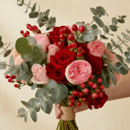 Hand-tied bouquet of red and pink roses with red berries and eucalyptus, held against a beige background.