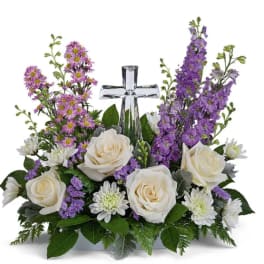 White roses and purple flowers arranged around a clear glass cross.
