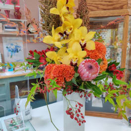 Tall arrangement with yellow orchids, orange blooms, pink protea and red berries in a white rectangular vase.