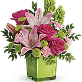 Pink lilies and roses with green accents arranged in a green cube vase