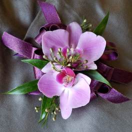 Lavender orchid corsage with small buds and dark purple ribbon on gray fabric.