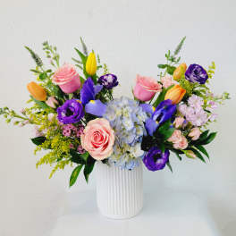 Mixed bouquet of pink roses, purple blooms, and a blue hydrangea in a white vase