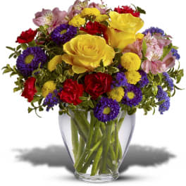 Bright mixed bouquet with yellow roses, red carnations, and purple blooms in a clear glass vase