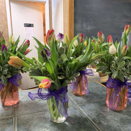 Four arrangements of mixed-color tulips in small glass vases with purple ribbon bows.