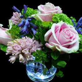 Short arrangement of pink roses, hyacinths, and green blooms in a clear glass vase on a black background