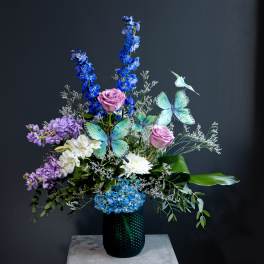 Tall floral arrangement with blue, purple, and white flowers in a dark vase