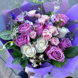 Hand-tied bouquet of lavender, pink, and white roses with accent flowers wrapped in purple paper.