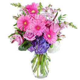 Mixed bouquet of pink gerbera daisies, lavender roses, and purple hydrangeas in a clear glass vase