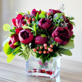 Deep pink peonies arranged in a clear square vase with berries and greenery.