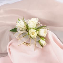 White rose wrist corsage with gold ribbon on soft pink fabric.