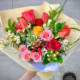 Hand-tied bouquet of mixed roses, tulips, and small white flowers wrapped in pale yellow paper