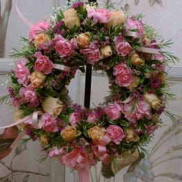 Round wreath of pink and peach roses with small purple blossoms and pink ribbons on a stand