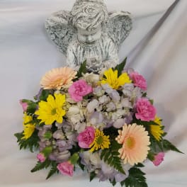 Angel figurine with low pastel flower arrangement of yellow daisies, pink blooms, and hydrangeas at the base.