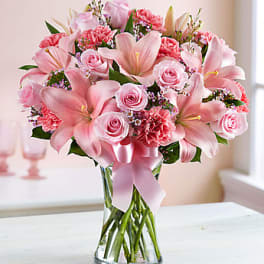 Pink lilies and roses arranged in a clear glass vase with a pink ribbon