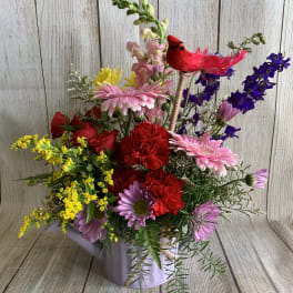 Colorful mixed flower arrangement in a lavender container with a red bird pick