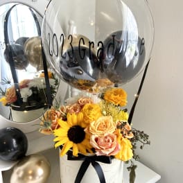 Graduation balloon arrangement with yellow and peach flowers in a hatbox