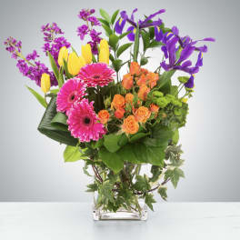 Mixed bouquet with pink gerbera daisies, orange roses, yellow tulips, and purple irises in a glass vase