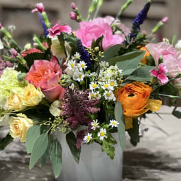 Colorful mixed arrangement of pink roses, yellow blooms, daisies and orange ranunculus in a white vase