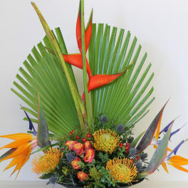 Tall tropical arrangement with orange and yellow flowers in a black shallow bowl.