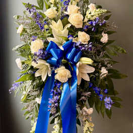 Standing spray of white roses and lilies with blue flowers and a large blue bow on an easel.