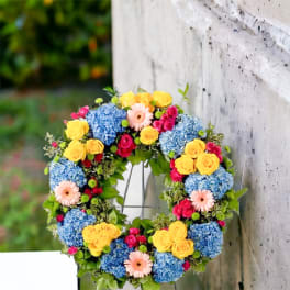 Bright floral wreath with blue hydrangeas, yellow roses, and pink blooms on a metal stand.