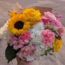 Round mixed bouquet with sunflower, yellow roses, pink carnations, and white hydrangeas in a clear vase