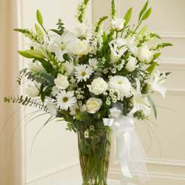 White lilies and daisies arranged in a tall glass vase with a white ribbon