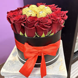 Red roses arranged in a black round box with gold-wrapped chocolates and a red ribbon.