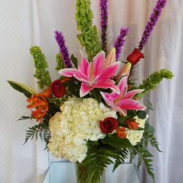Tall bouquet with pink lilies, red roses, white hydrangeas, and purple spikes in a glass vase