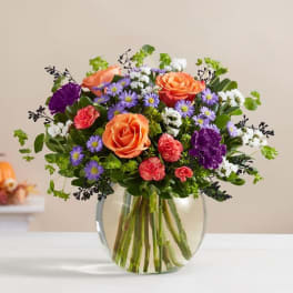 Mixed bouquet of orange roses, purple asters, and carnations in a glass vase