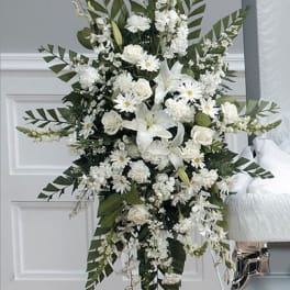 Tall all-white standing spray of lilies, roses, carnations, and daisies on an easel beside a casket