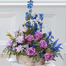 Basket arrangement of purple roses, lavender daisies, and blue flowers with tall blue accents