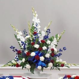 Tall red, white, and blue funeral arrangement with roses, carnations, and blue delphinium in a white pedestal vase