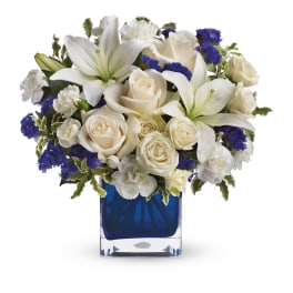 Compact arrangement of white lilies, roses, and carnations with blue accents in a blue glass cube vase