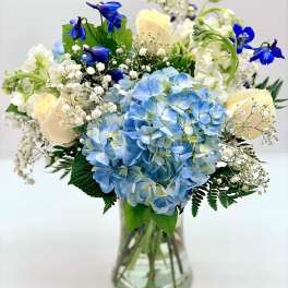 Vase arrangement with blue hydrangeas, cream roses, white blooms, and bright blue flowers