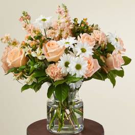 Peach roses, white daisies, and pastel blooms arranged in a clear glass vase.