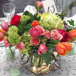 Low centerpiece of pink, orange, and red blooms in a square glass vase on a banquet table