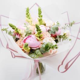 Hand-tied bouquet of pink peonies, peach roses, and snapdragons wrapped in white paper in a glass vase