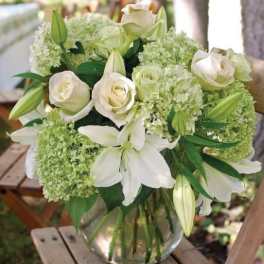 White lilies, roses, and green hydrangeas in a clear glass vase on a wooden stool outdoors