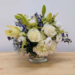 White roses, lilies, and hydrangeas with blue accents arranged in a low clear glass vase