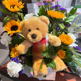 Stuffed teddy bear nestled among yellow sunflowers, white carnations, and pink and purple daisies.