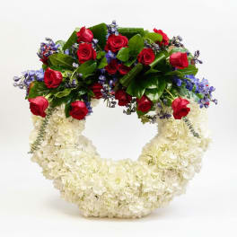 Round wreath of white flowers topped with red roses and blue accent blooms