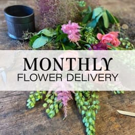 Loose flowers and greenery on a table with a black cup and monthly flower delivery text