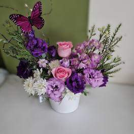 Purple and pink flower arrangement in a white pot with a butterfly pick