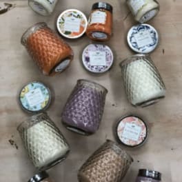 Assorted colored candles in textured glass jars with decorative lids arranged on a wooden surface