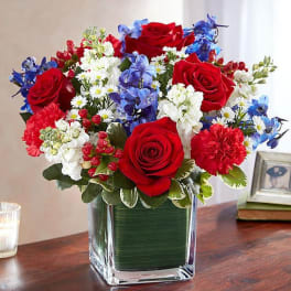 Red roses, blue flowers, and white daisies arranged in a square glass vase